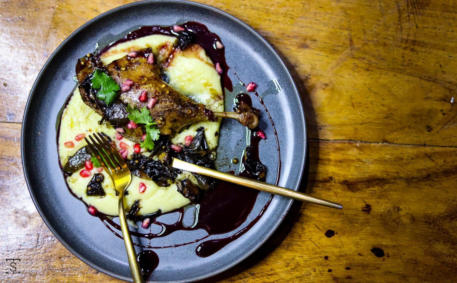 Roasted leg on silky purée with a dark berry jus, pomegranate and gold cutlery on a rustic table